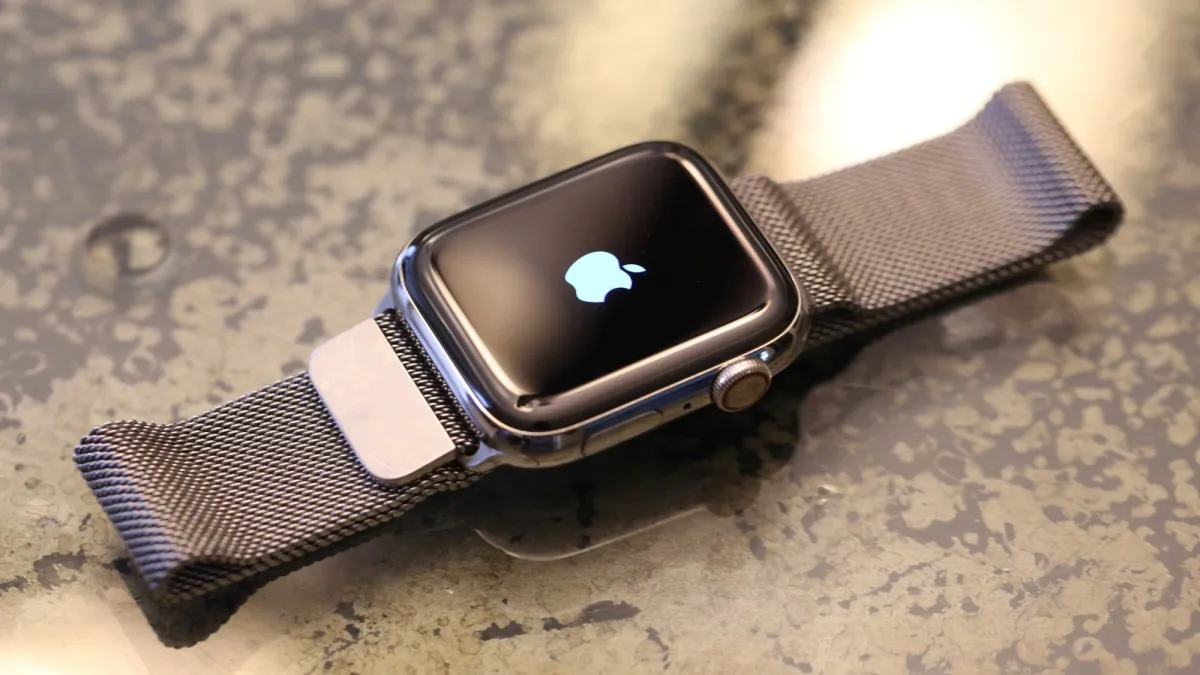 Apple Watch Series terbaru dengan tali sport biru muda dipajang di atas meja kayu, menunjukkan layar cerah yang menampilkan waktu dan tanggal saat ini beserta ikon aplikasi kesehatan.