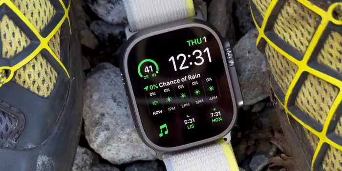 Close-up Apple Watch dengan casing stainless steel dan tali kulit coklat, terletak di samping iPhone, keduanya menampilkan wallpaper yang serasi dan notifikasi sinkronisasi panggilan masuk.